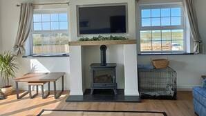 A living room with a fireplace, a TV mounted above it, and a window with curtains. - Aberporth 2-bed Cottage , Sleeps 4 & Pet Friendly (Cardigan)