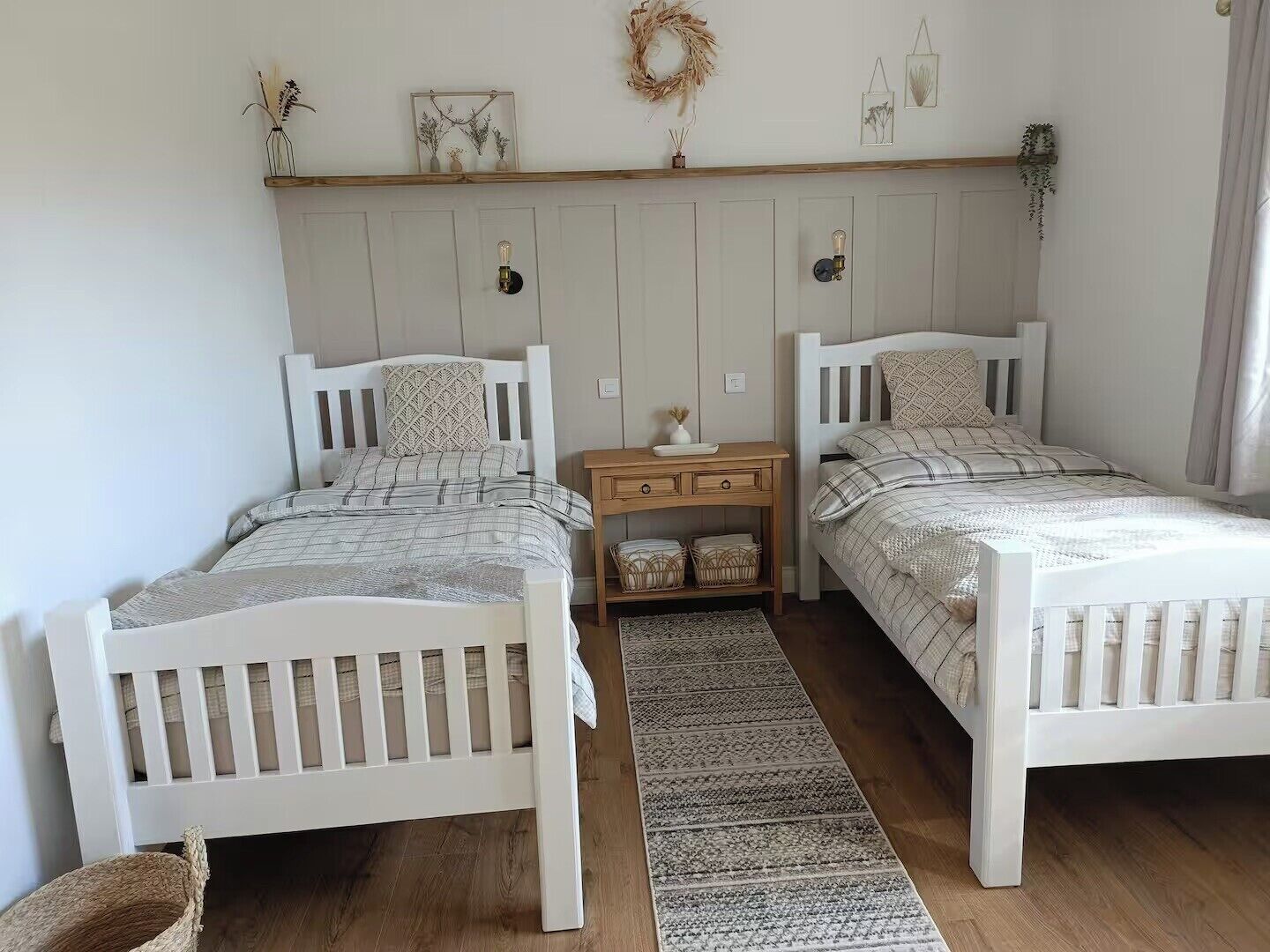 A bedroom with two white beds, a wooden nightstand, a woven rug, and a wall-mounted shelf with decorative items.