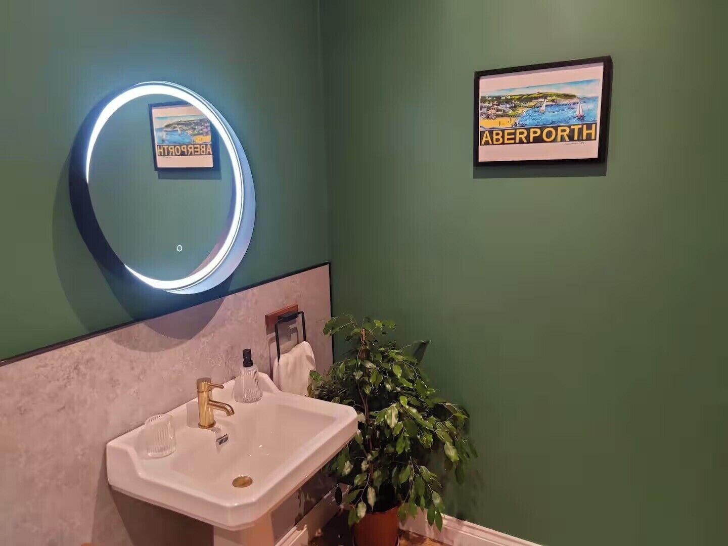 A bathroom with a sink, a mirror with a light, a framed picture of a coastal scene, and a green wall.