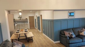 A modern living room with a grey sofa, a dining table set with plates, and a kitchen in the background. - Aberporth 2-bed Cottage , Sleeps 4 & Pet Friendly (Cardigan)