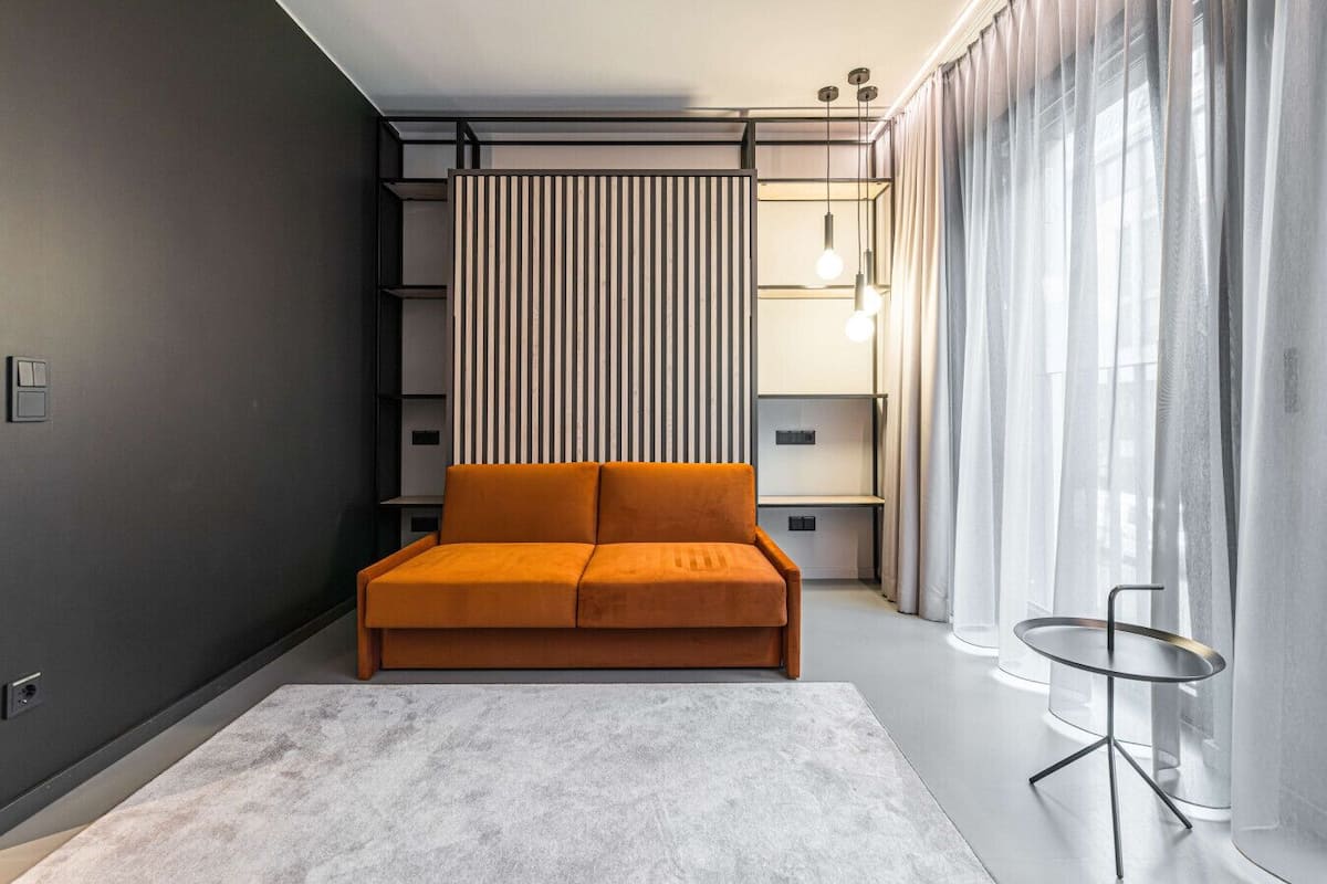 A modern bedroom with a sofa bed, a side table, a built-in shelving unit, and a large window with sheer curtains.