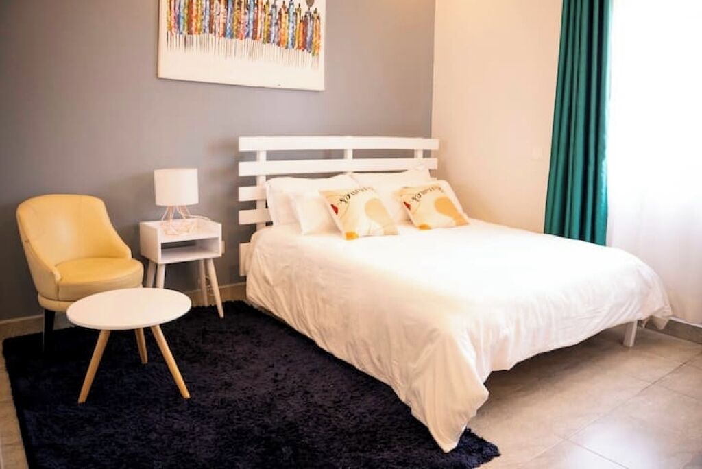 A bedroom with a white bed, a small bedside table with a lamp, a chair, and a dark rug.