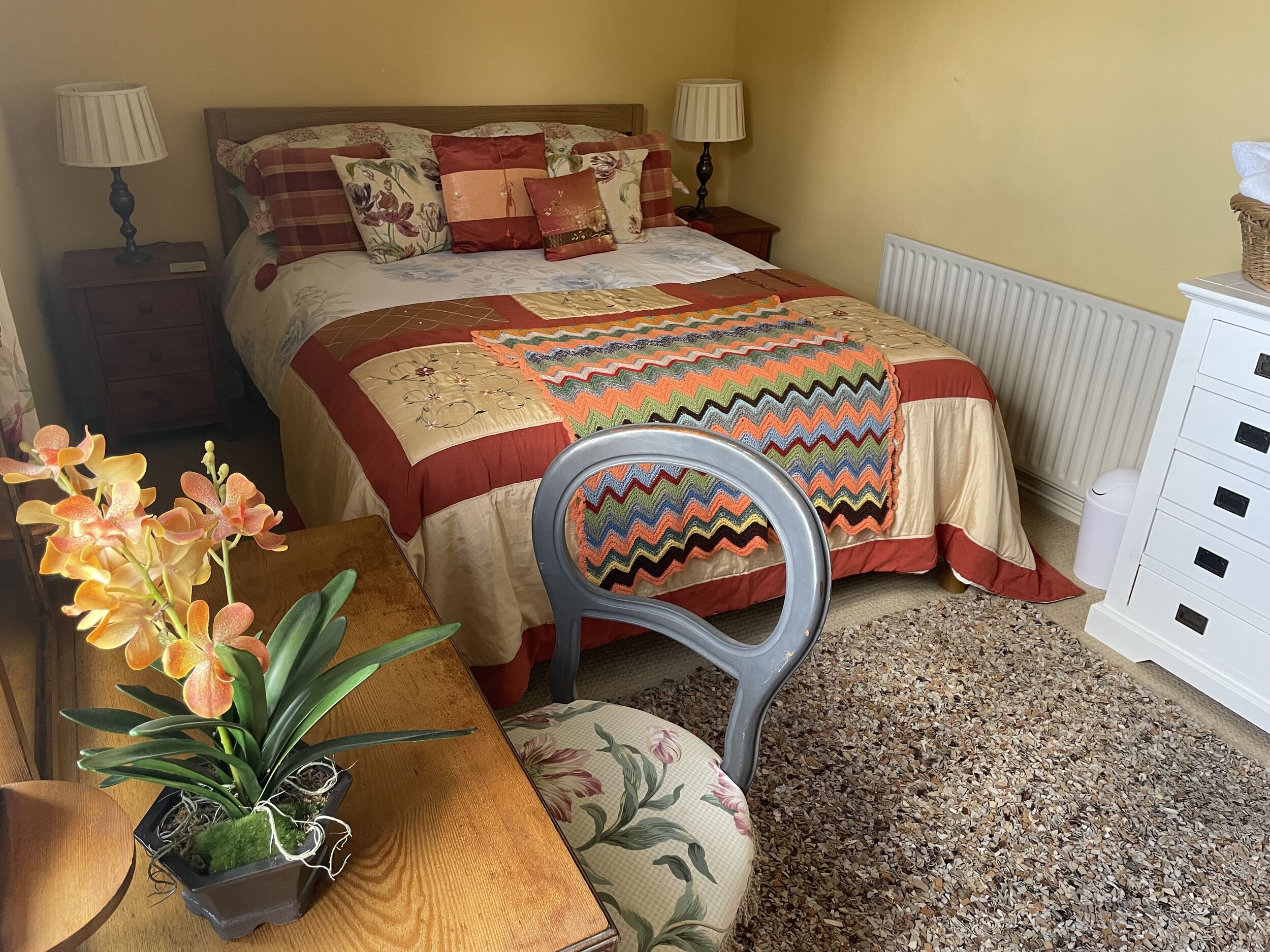 A bedroom with a bed, bedside tables, lamps, a chair, a small table with a plant, and a radiator.
