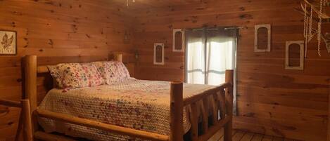 Una habitación de cabaña de madera con una cama, una ventana con cortinas y una lámpara colgante.