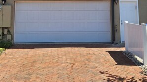 A two-story house with a garage, a window with blinds, and a brick driveway.