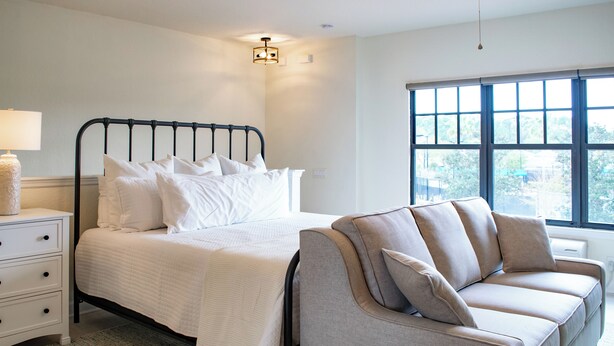 A bedroom with a bed, a nightstand, a lamp, a sofa, and a large window.
