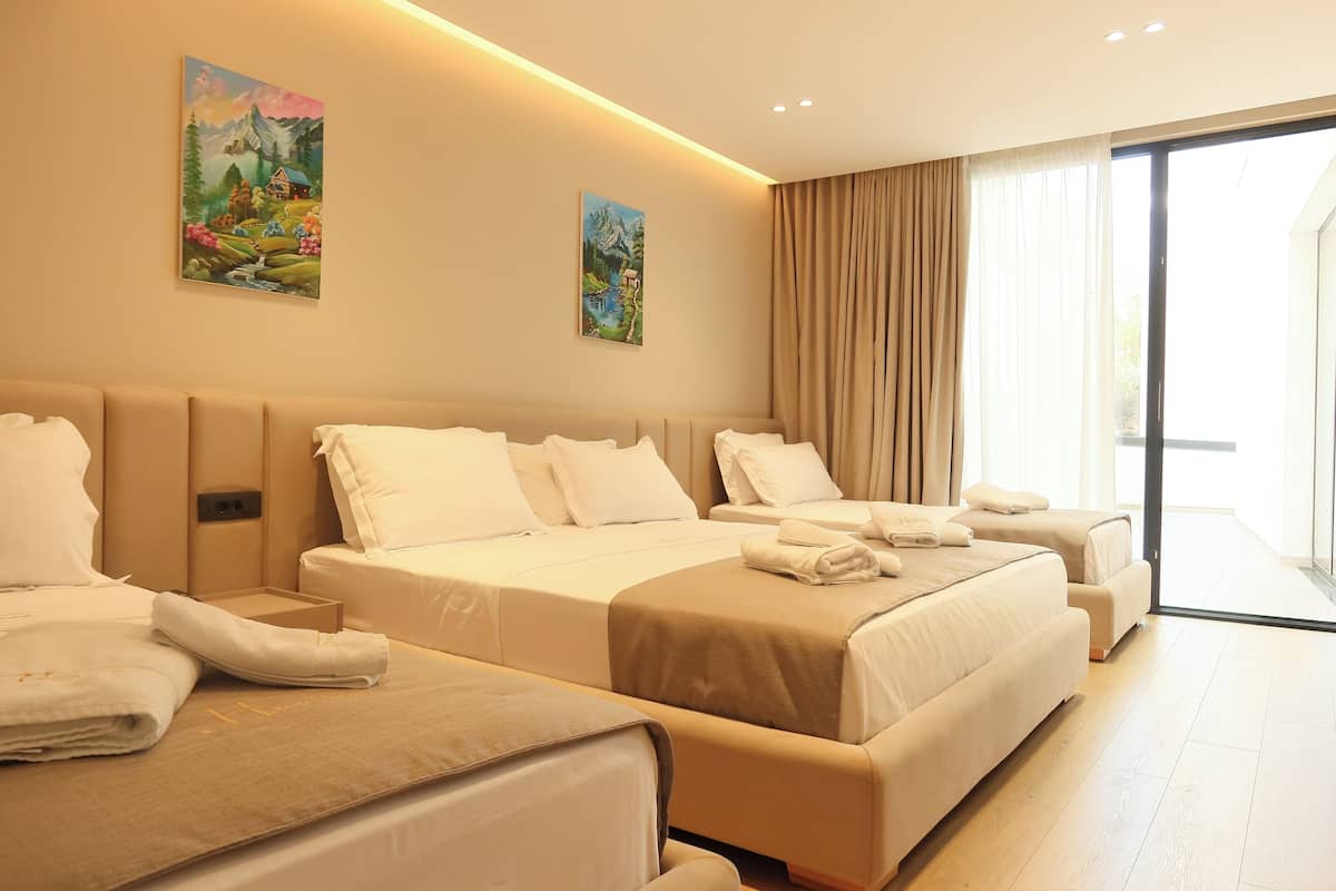 A hotel room with a large bed, a smaller bed, and a sofa. There are two paintings on the wall, a window with curtains, and a sliding glass door.