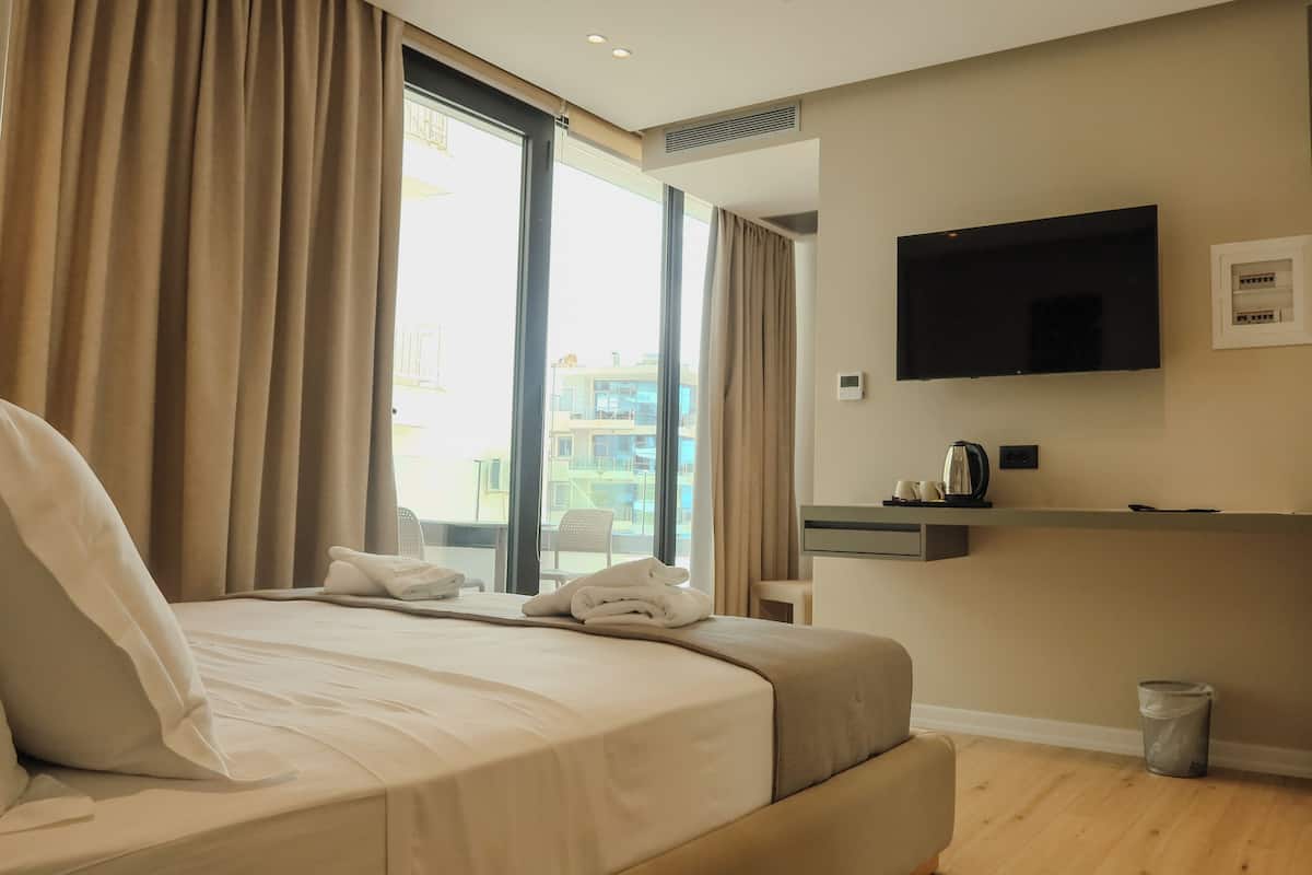 A hotel room with a bed, a TV mounted on the wall, a small table with a kettle, and a view of the outside.