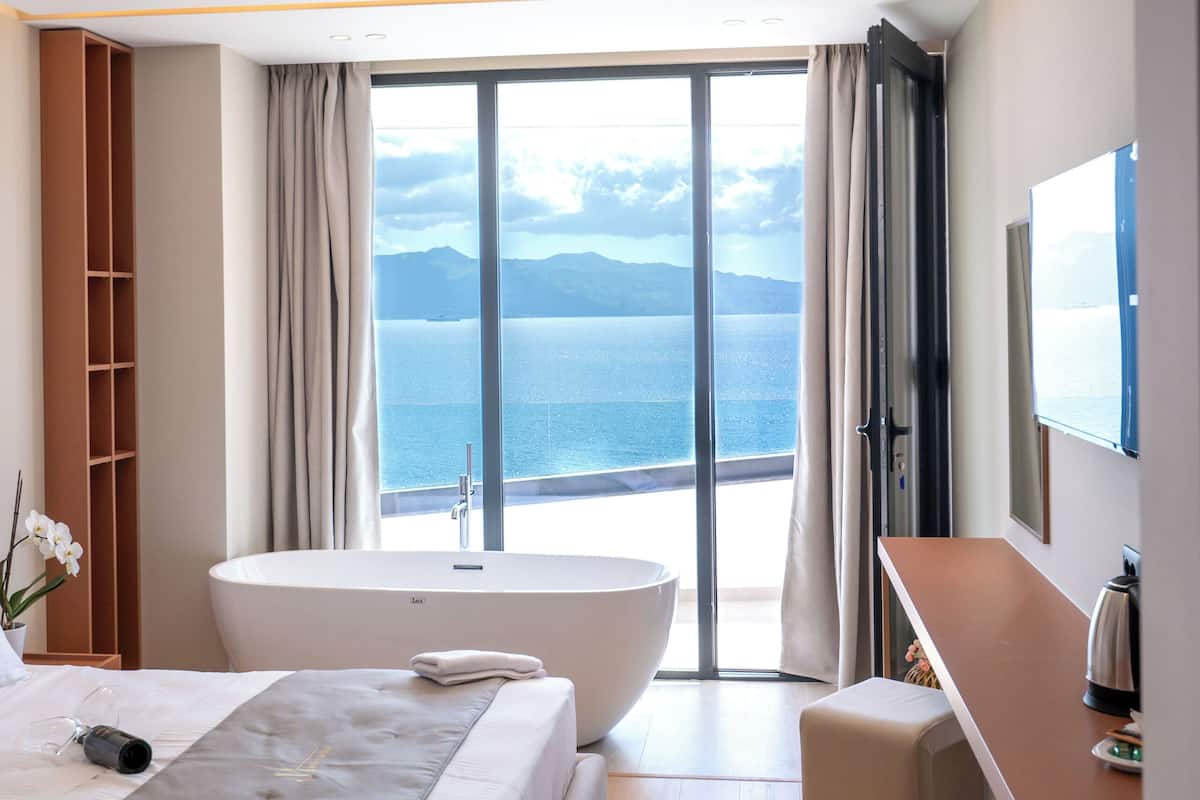 A modern hotel room with a large bed, a bathtub, and a view of the sea.
