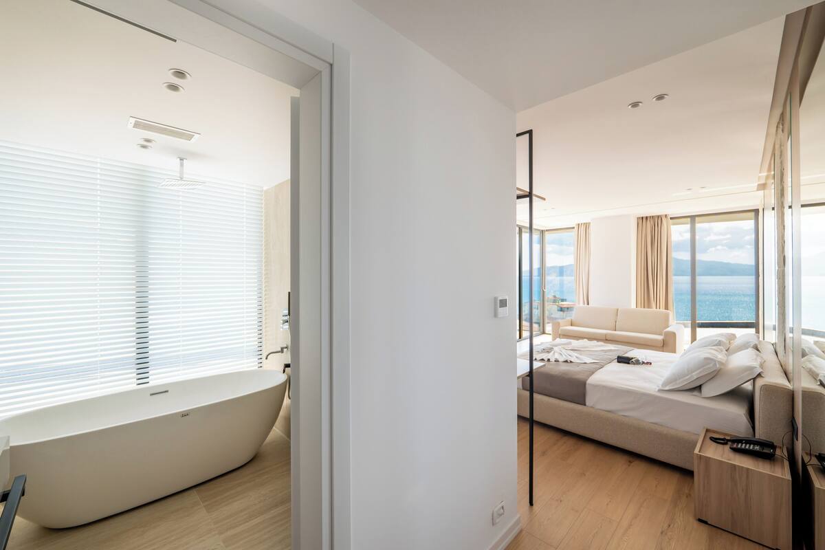 A modern hotel room with a large window, a bathtub, a sofa bed, and a wooden bedside table.