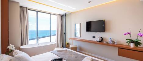 A modern hotel room with a large bed, a desk, a television, and a view of the sea.