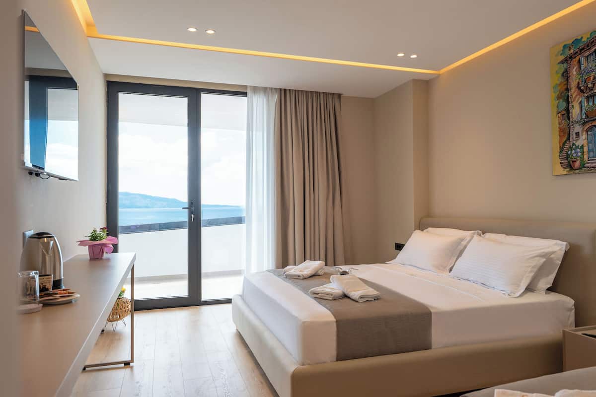 A modern hotel room with a large bed, a desk with a coffee maker, and a view of the sea through the sliding glass door.
