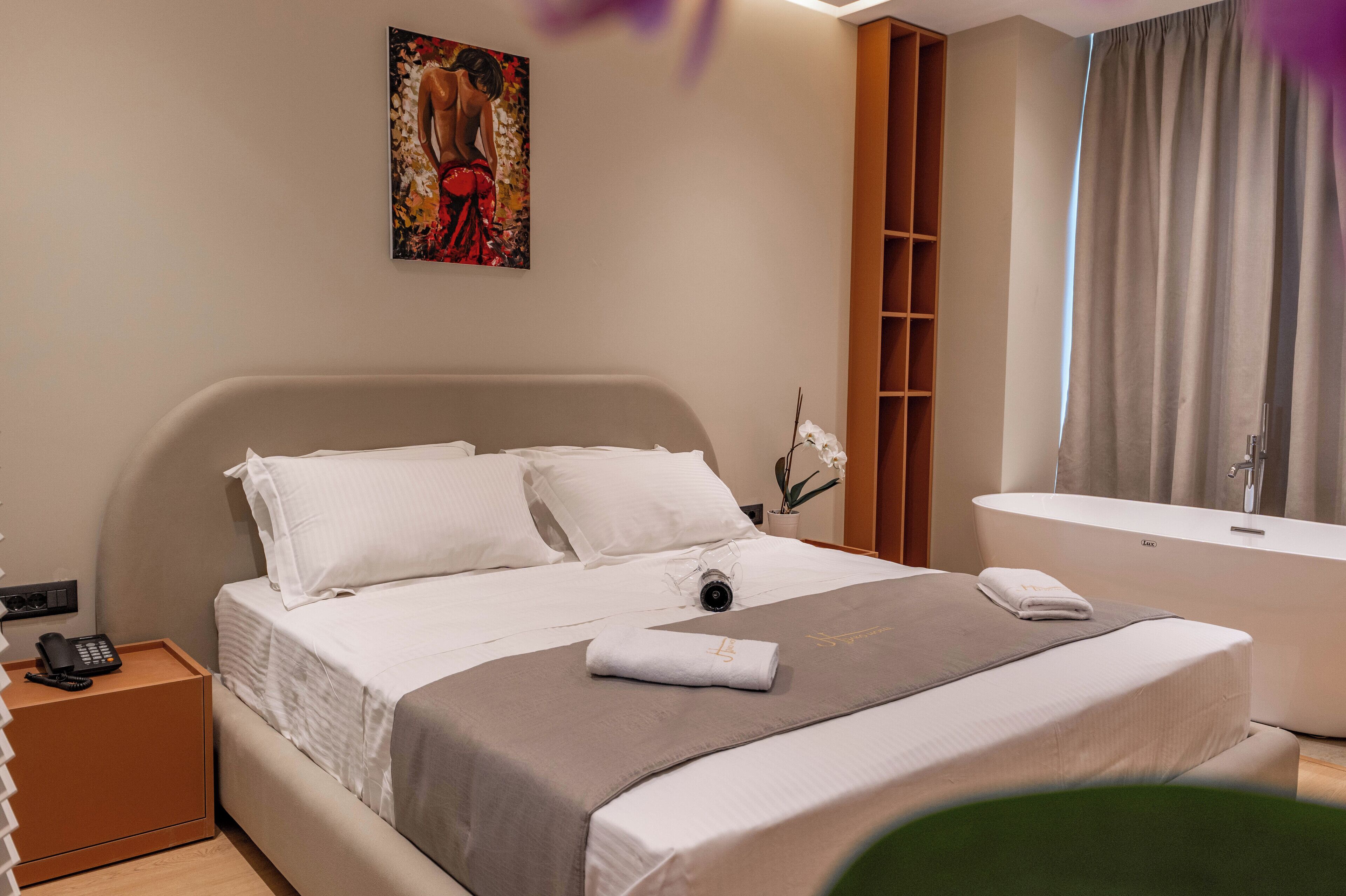 A hotel room with a bed, a telephone, a painting on the wall, a bathroom with a bathtub, and a curtain.