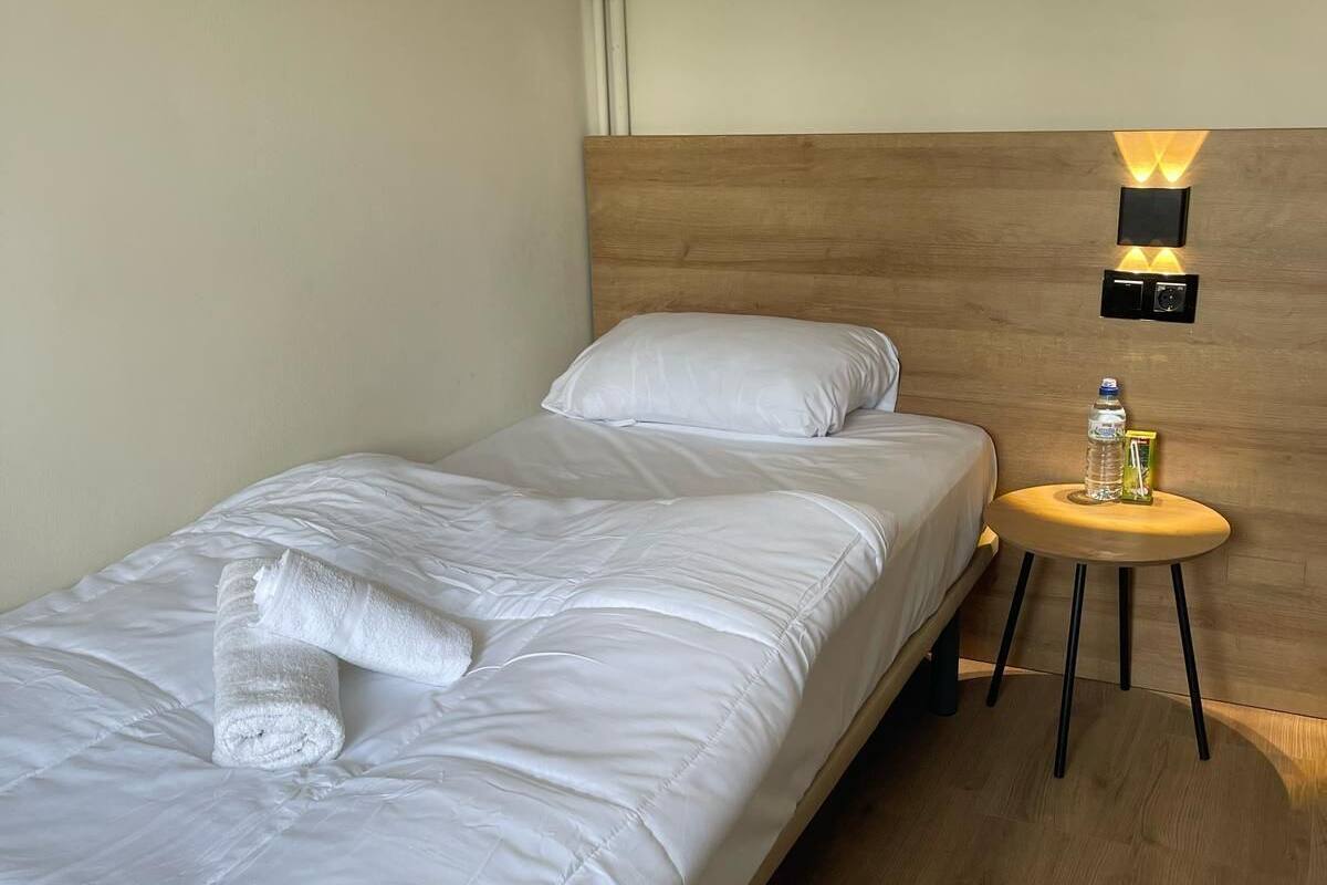 Una cama bien hecha con sábanas blancas y una toalla enrollada, una pequeña mesa redonda de madera con una botella y un vaso, y una lámpara fijada a la pared.
