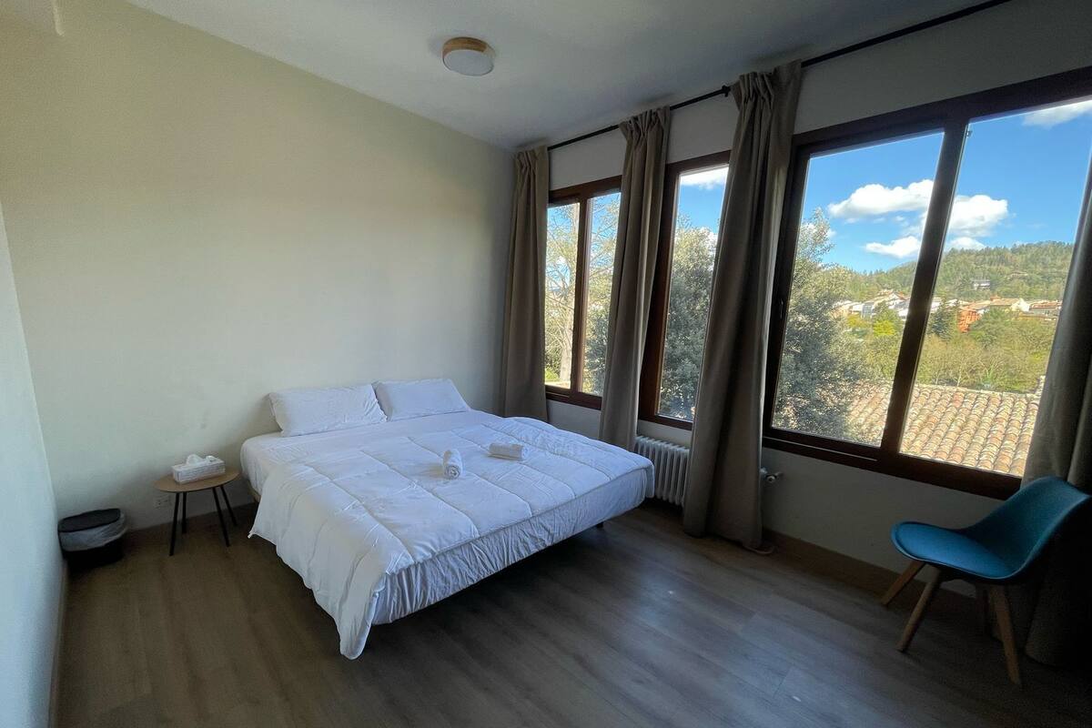 Un dormitorio con una cama grande, una mesita pequeña, una silla y vistas a un pueblo a través de amplios ventanales.