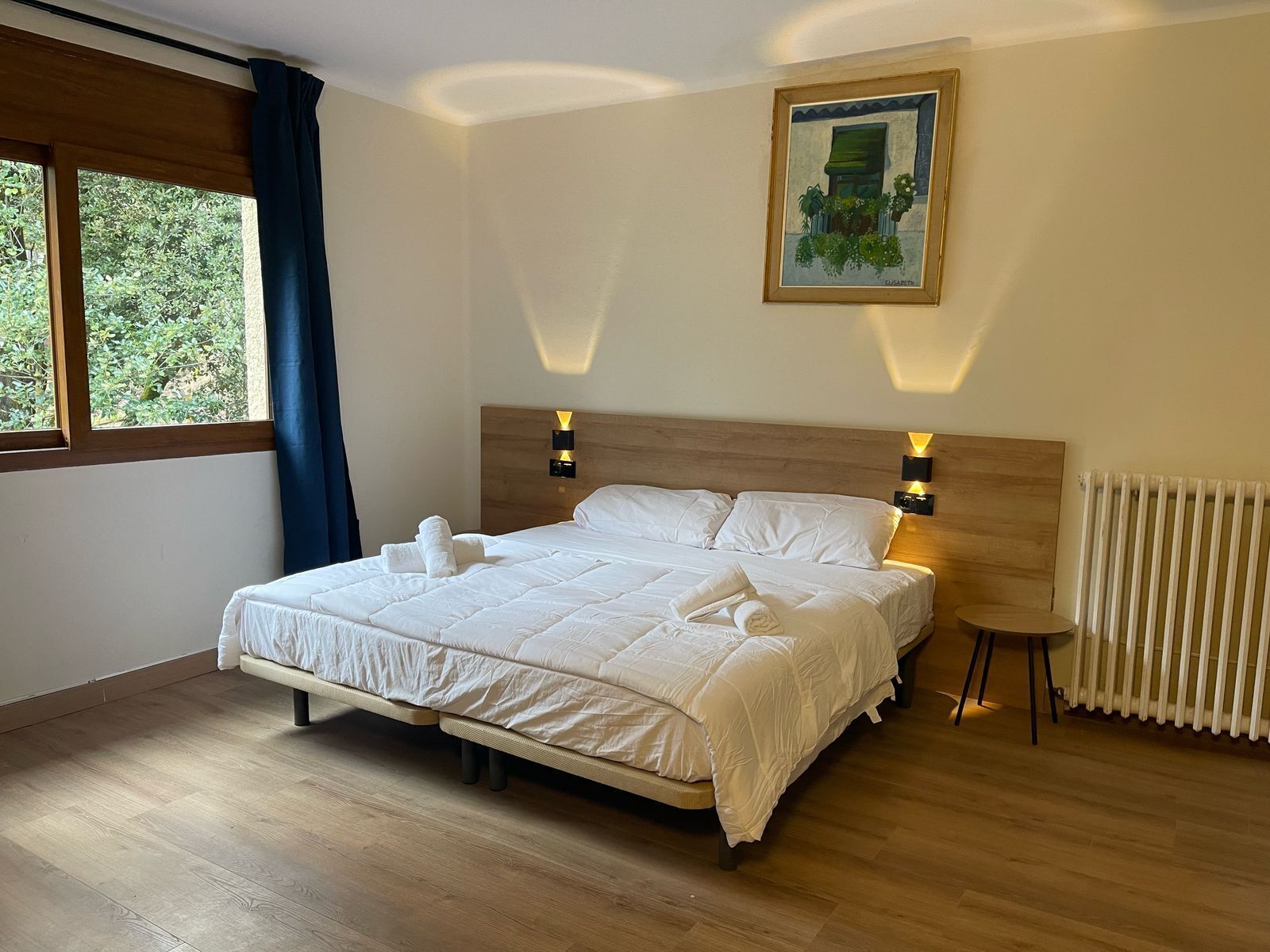 A neatly made double bed with white linens, two bedside lamps, a small bedside table, and a painting on the wall.