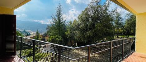 Un balcón con barandilla metálica curva, piso de baldosas y vista a un paisaje montañoso con árboles y edificios a lo lejos.