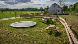 Eine ländliche Außenanlage mit Holzpferd, Trampolin, Picknicktischen und einem Haus mit rundem Fenster.