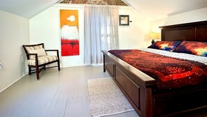 Un dormitorio con techo inclinado, una cama con ropa de cama estampada, una silla, un cuadro y una ventana con cortinas.