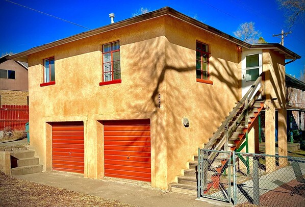 Un edificio de dos pisos con una puerta de garaje roja, una escalera que conduce al nivel superior y un pequeño porche.