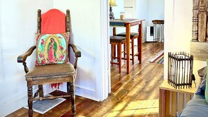Una habitación con piso de madera, una silla con un cojín de colores, una mesita y una cama con cabecera de madera.