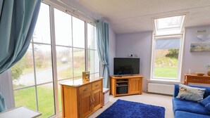 Una sala de estar con un televisor sobre un mueble de madera, un sofá azul, una mesita y ventanas amplias con cortinas.
