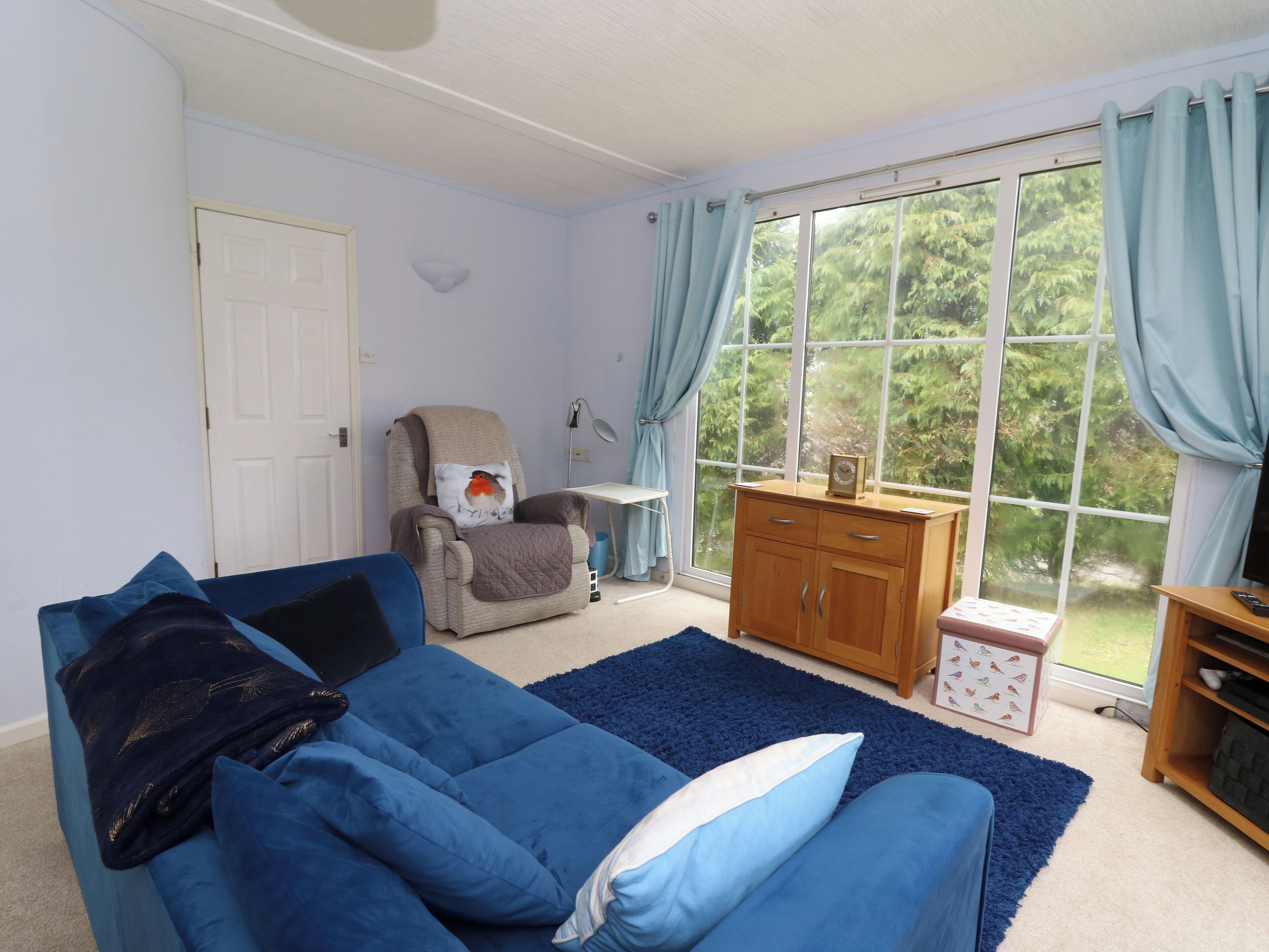 Uma sala de estar com um sofá azul, uma poltrona com almofada, um armário de madeira e uma janela grande com cortinas.