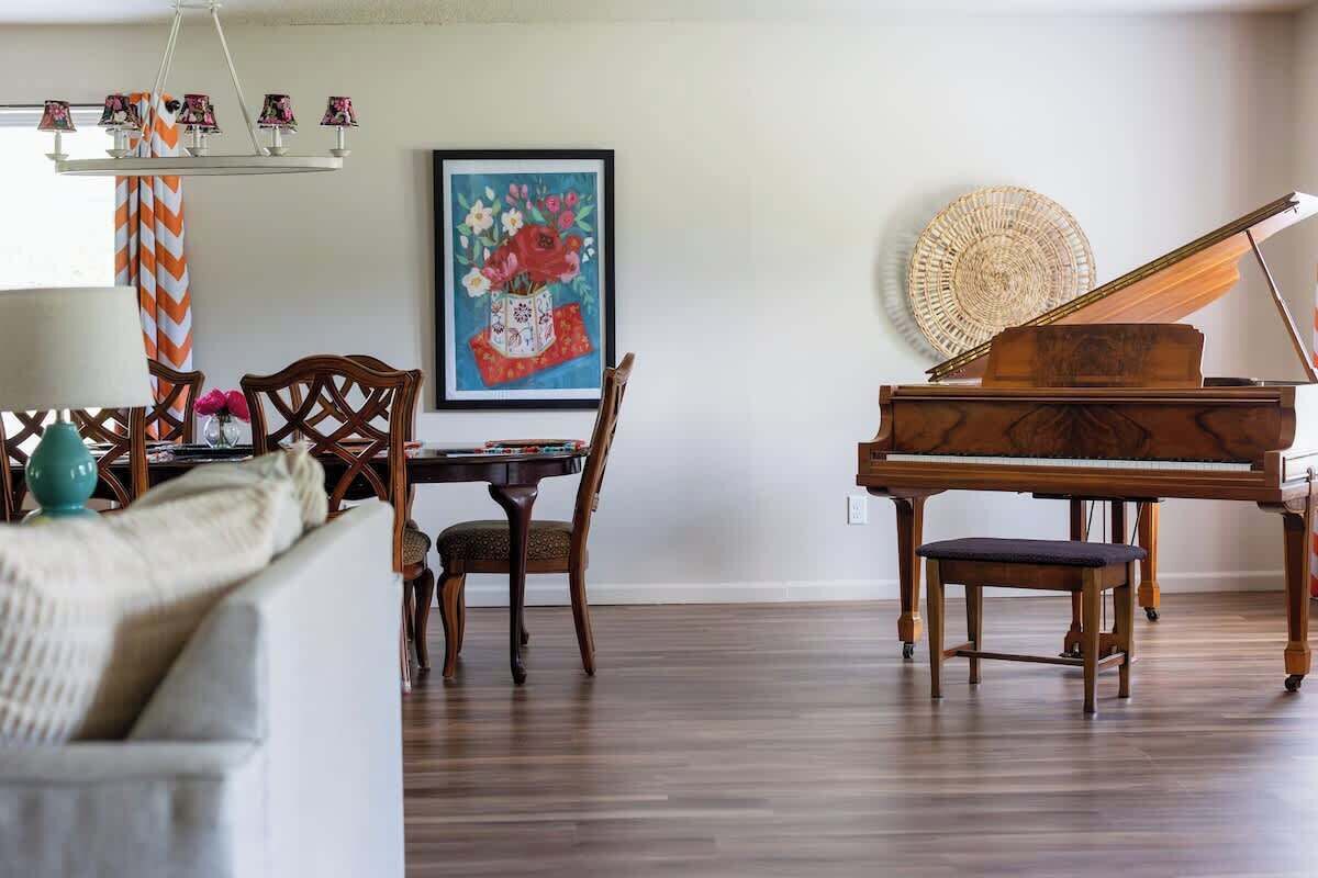 A living room with a wooden piano, a dining table with chairs, a sofa, and a framed painting on the wall.