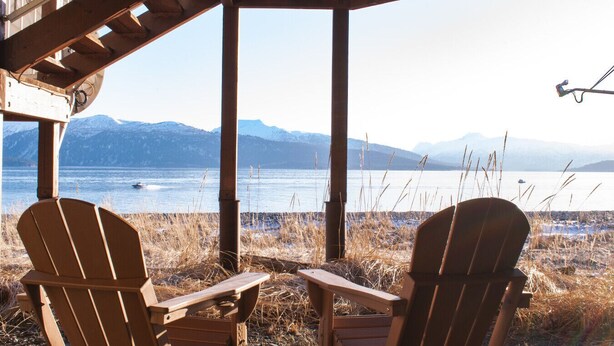 Två Adirondack-stolar placerade framför en sjö, med berg i bakgrunden, under en träkonstruktion.