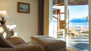 A room with a sofa, a lamp, a framed picture on the wall, and a large window offering a view of a mountainous landscape. - Lakeshore Lodging Ocean Front Lodges (Homer)