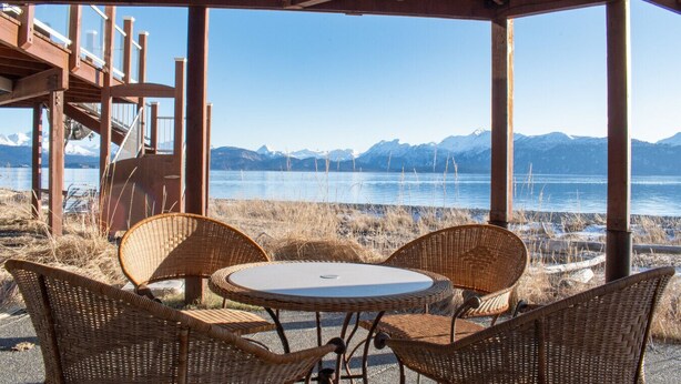 A covered outdoor seating area with wicker chairs and a table, overlooking a lake with mountains in the background. - Lakeshore Lodging Ocean Front Lodges (Homer)