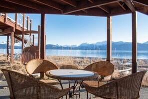 Ein überdachter Außenbereich mit Korbstühlen und einem Tisch, mit Blick auf einen See und Berge im Hintergrund.
