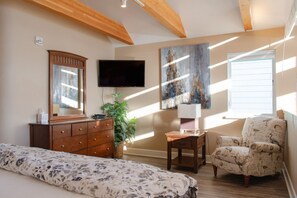 A bedroom with a bed, dresser, TV, artwork, chair, and a window. - Lakeshore Lodging Ocean Front Lodges (Homer)