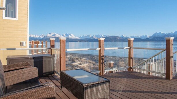 A wooden deck with outdoor furniture, a glass table, and a view of a lake with mountains in the background. - Lakeshore Lodging Lodge 713 (Homer)