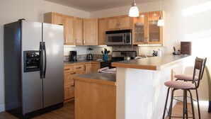 A modern kitchen with wooden cabinets, a stainless steel refrigerator, a microwave, and a coffee maker. - Lakeshore Lodging Lodge 713 (Homer)
