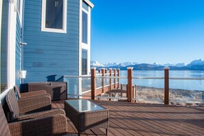 A deck with wicker furniture, a glass table, and a view of a lake with mountains in the background. - Lakeshore Lodging Lodge 715 (Homer)