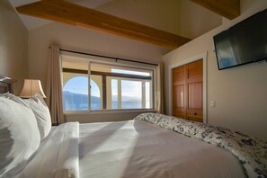 A bedroom with a bed, a window with curtains, a TV mounted on the wall, and a wooden ceiling beam.