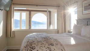 A bedroom with a bed, a television, a window with curtains, and a view of the sea. - Lakeshore Lodging Lodge 715 (Homer)