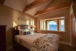 A bedroom with a bed, a window with a view, a wooden ceiling, and a wall-mounted lamp.
