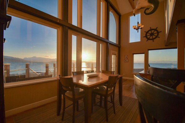 A dining area with a view of the sea, a piano, and a ship's wheel decoration.