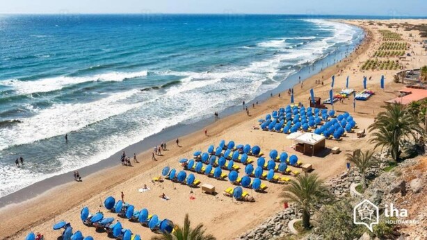 Ein Strand mit Reihen blauer Sonnenschirme und Liegestühlen, einem klaren Himmel und dem Meer.