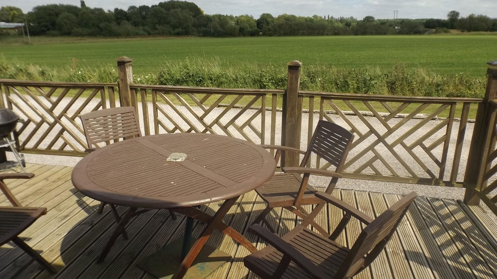 An outdoor deck with a round table and four chairs, surrounded by a wooden fence with a view of a green field.