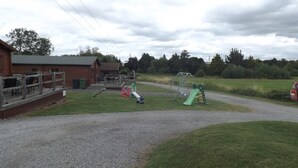 A playground with a slide and swings, a gravel path, and a building with a fenced area. - Two Bedroom Timber Lodge, Avonvale Holiday Lodges (Evesham)