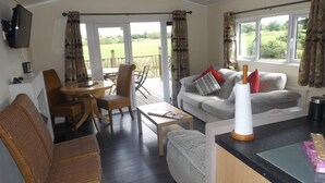 A cozy living room with a sofa, a coffee table, and a dining area with chairs. There are large windows with curtains, a flat-screen TV mounted on the wall, and a view of a green landscape outside. - Two Bedroom Timber Lodge, Avonvale Holiday Lodges (Evesham)
