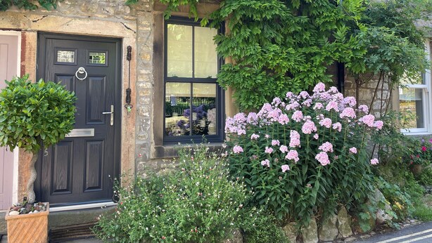 Ein Steinhaus mit einer dunklen Tür, einem Fenster mit Spiegelung und rosa Blumen davor.
