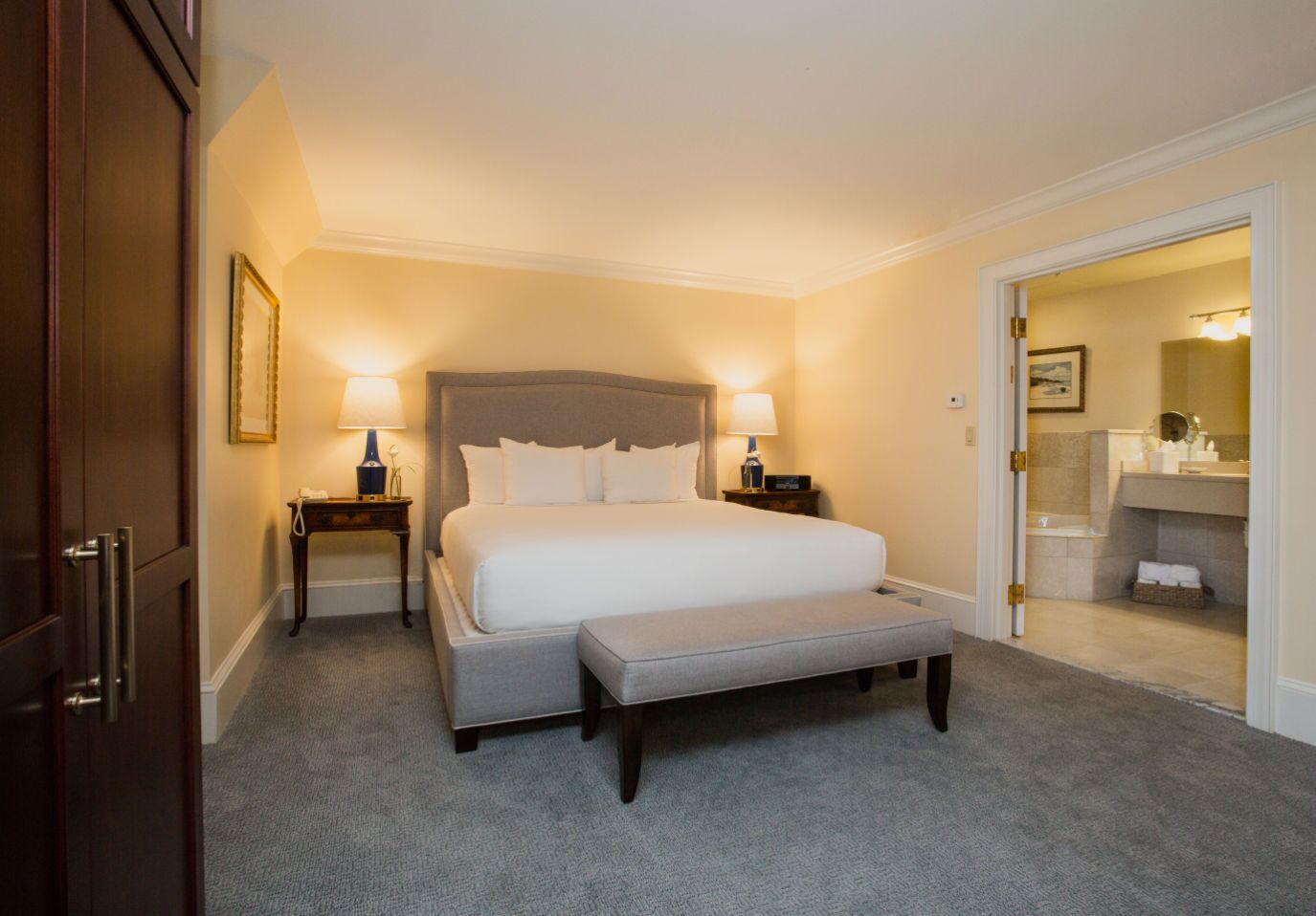 Camera d'albergo con un letto grande, comodini con lampade, una panca e un bagno visibile attraverso una porta aperta.
