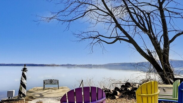 Blick auf einen See mit einem Steg, einer Bank und bunten Adirondack-Stühlen.