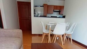 Una sala moderna con una mesa de centro de madera, un sofá beige y un comedor con sillas blancas, con una cocina al fondo.