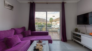 A living room with a purple sofa, a coffee table with a bowl of pasta, a TV on the wall, and a balcony with a view of the mountains. - Beachfront Apartment in Playa de Mogan (Mogan)