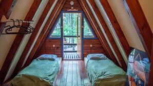 A cozy attic bedroom with two beds, wooden beams, and a large window offering a view of the forest.